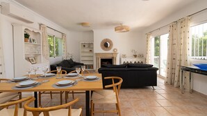 Un comedor con una mesa de madera dispuesta para cuatro personas, rodeada de sillas de madera. Se observa un sofá negro y una chimenea al fondo.