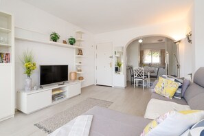 A modern living room with a flat-screen TV, white furniture, and a dining area visible through an archway.