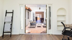 Une pièce avec un plancher en bois, un bureau avec une chaise, une échelle sur laquelle sont posées des serviettes, et une vue sur un coin repas avec des chaises et une table.