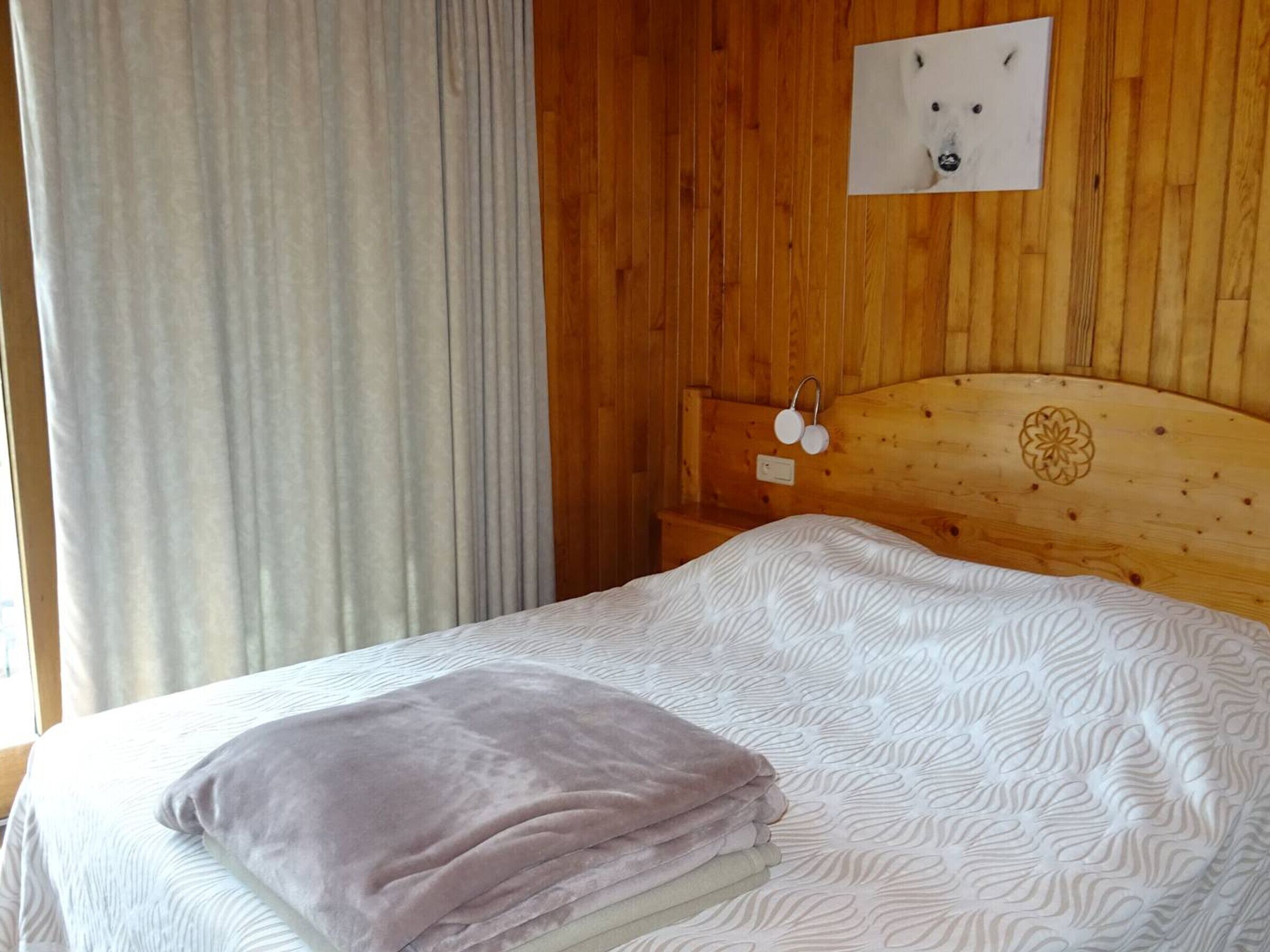 A wooden-paneled room with a bed, a headboard with a decorative emblem, a wall-mounted light switch, and a framed picture on the wall.