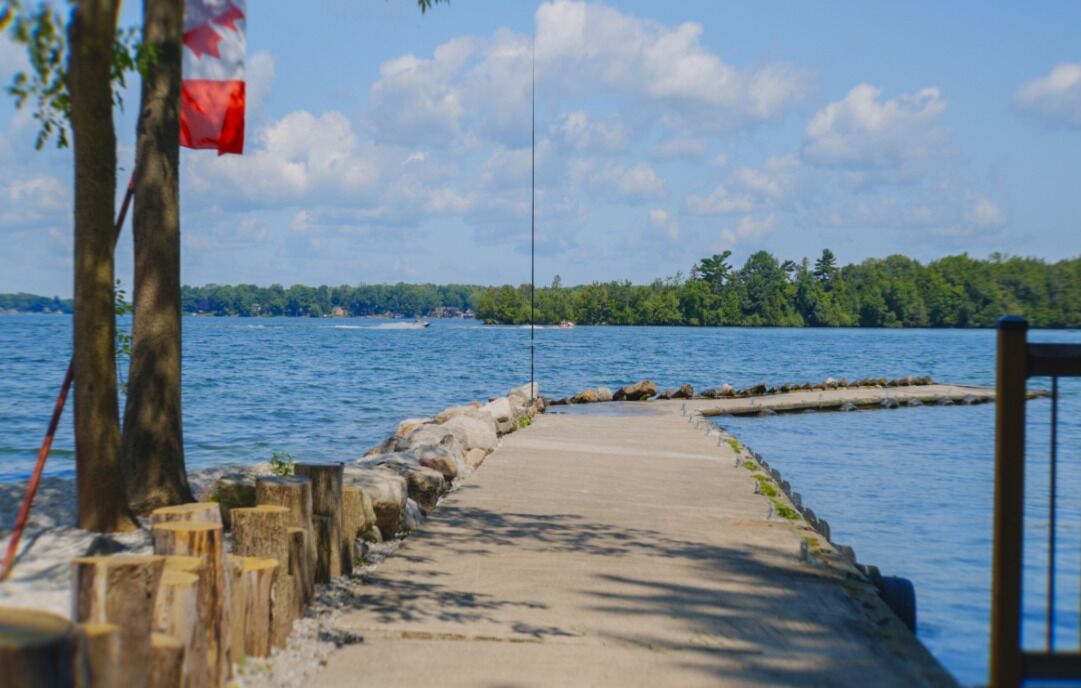 Un quai en béton s’étendant dans un lac, sur lequel flotte un drapeau canadien, le tout entouré d’arbres et sous un ciel dégagé.