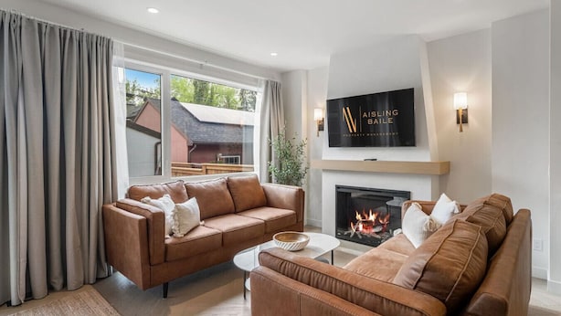 A modern living room with two brown leather sofas, a white coffee table, a fireplace, and a TV mounted above it. - La More | Hot Tub | Kensington | Luxury | Office (Calgary)