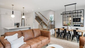 A modern living room with a brown leather sofa, a dining area with a wooden table and black chairs, and a kitchen with white cabinets and pendant lighting. - La More | Hot Tub | Kensington | Luxury | Office (Calgary)