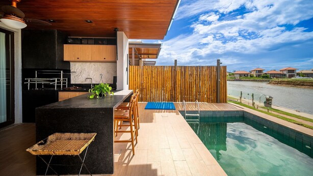 A modern kitchen with a wooden ceiling, a poolside area with a bamboo fence, and a view of houses across a water body.