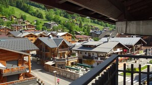 Ein Holzhaus mit Balkon und Außenbereich mit Sitzgelegenheiten, umgeben von ähnlichen Gebäuden und einer grünen Hügelkulisse im Hintergrund.