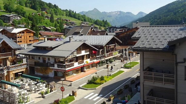 Ein Bergdorf mit traditionellen Holzhäusern, einer zentralen Straße mit Geschäften und Blick auf grüne Hügel und Berge im Hintergrund.