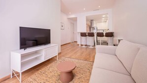 A modern living room with a flat-screen TV, a white entertainment unit, a beige sofa, and a round coffee table. There is a kitchen area in the background with bar stools.