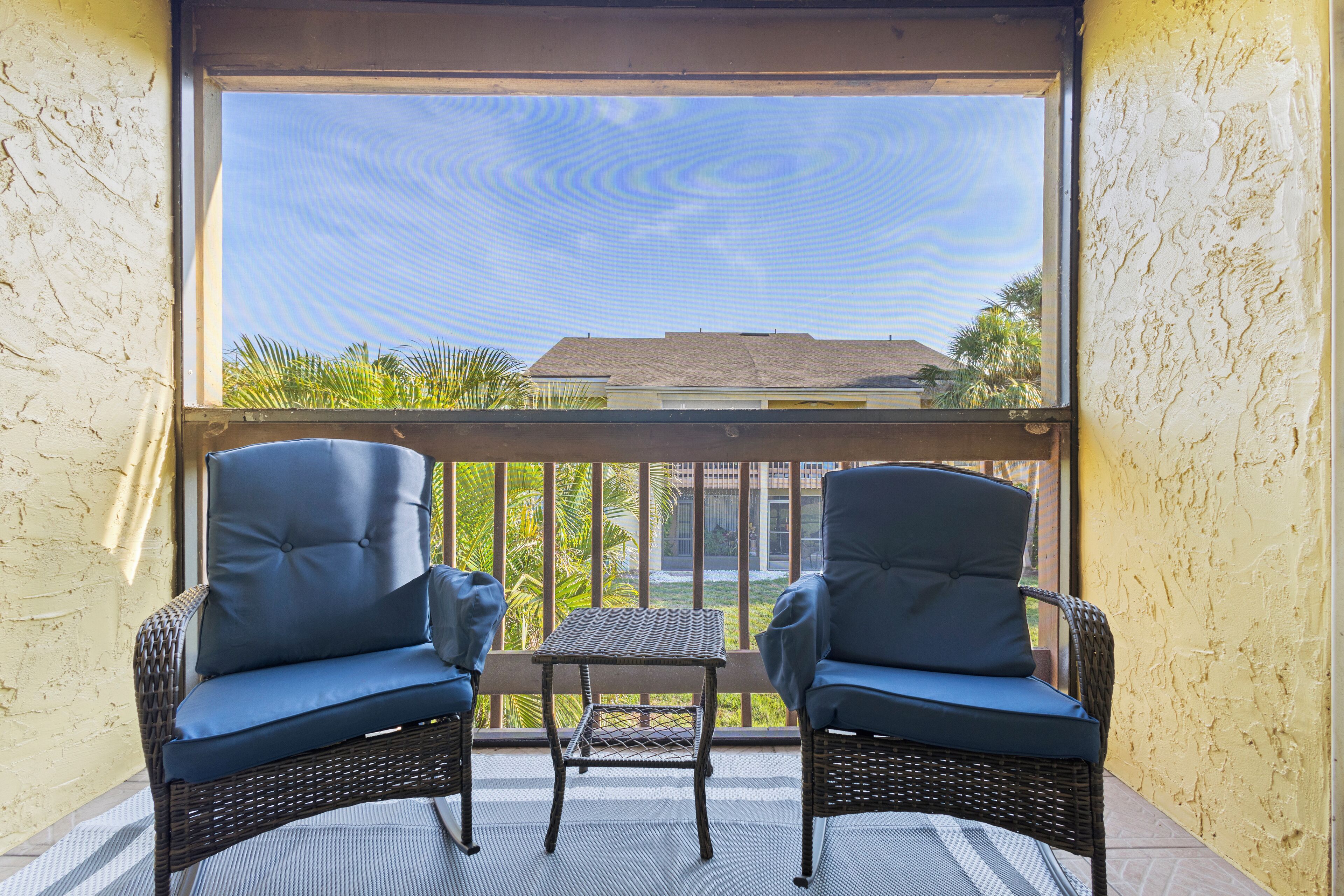 A balcony with wicker chairs and a small table, overlooking a building and palm trees.