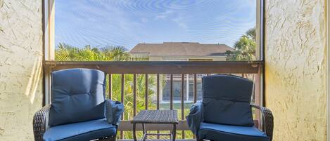 A balcony with wicker chairs and a small table, overlooking a building and palm trees.