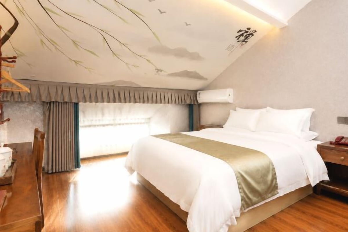 A hotel room with a large bed, a desk with a chair, a window with curtains, and a wall-mounted air conditioner.
