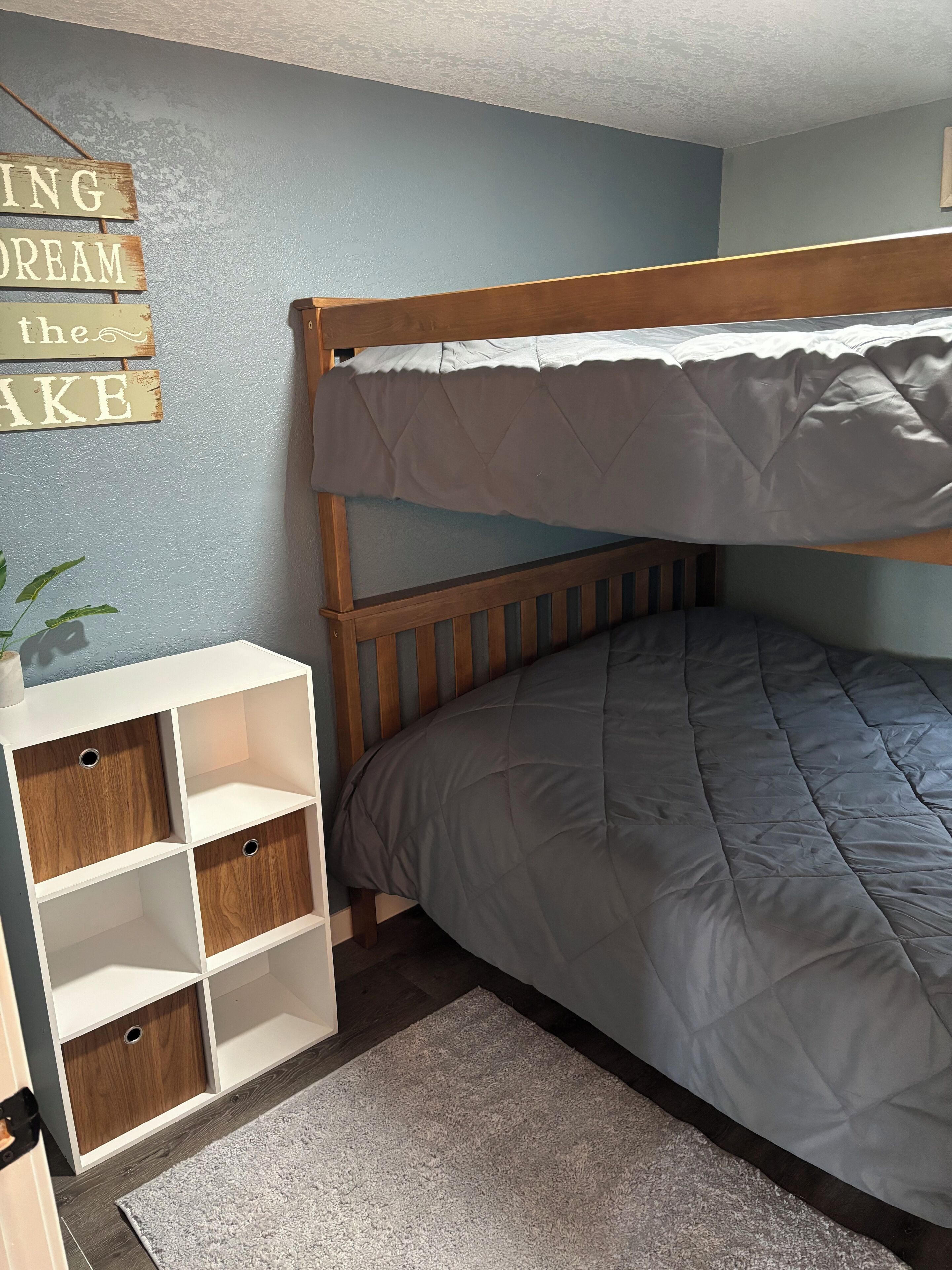 A bunk bed with a grey quilt, a white storage unit with wooden shelves, and a wall sign.