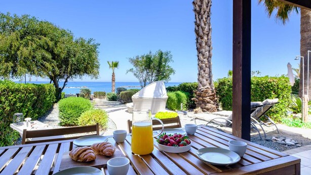 Ein Holztisch mit Croissants, einer Obstschale und einem Glas Orangensaft, mit Blick auf eine malerische Küstenlandschaft.