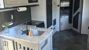 A compact kitchen with a built-in microwave, stove, and sink. There is a small island with a sink and a bottle of dish soap. The kitchen has a herringbone-patterned backsplash and dark gray tiles on the floor.