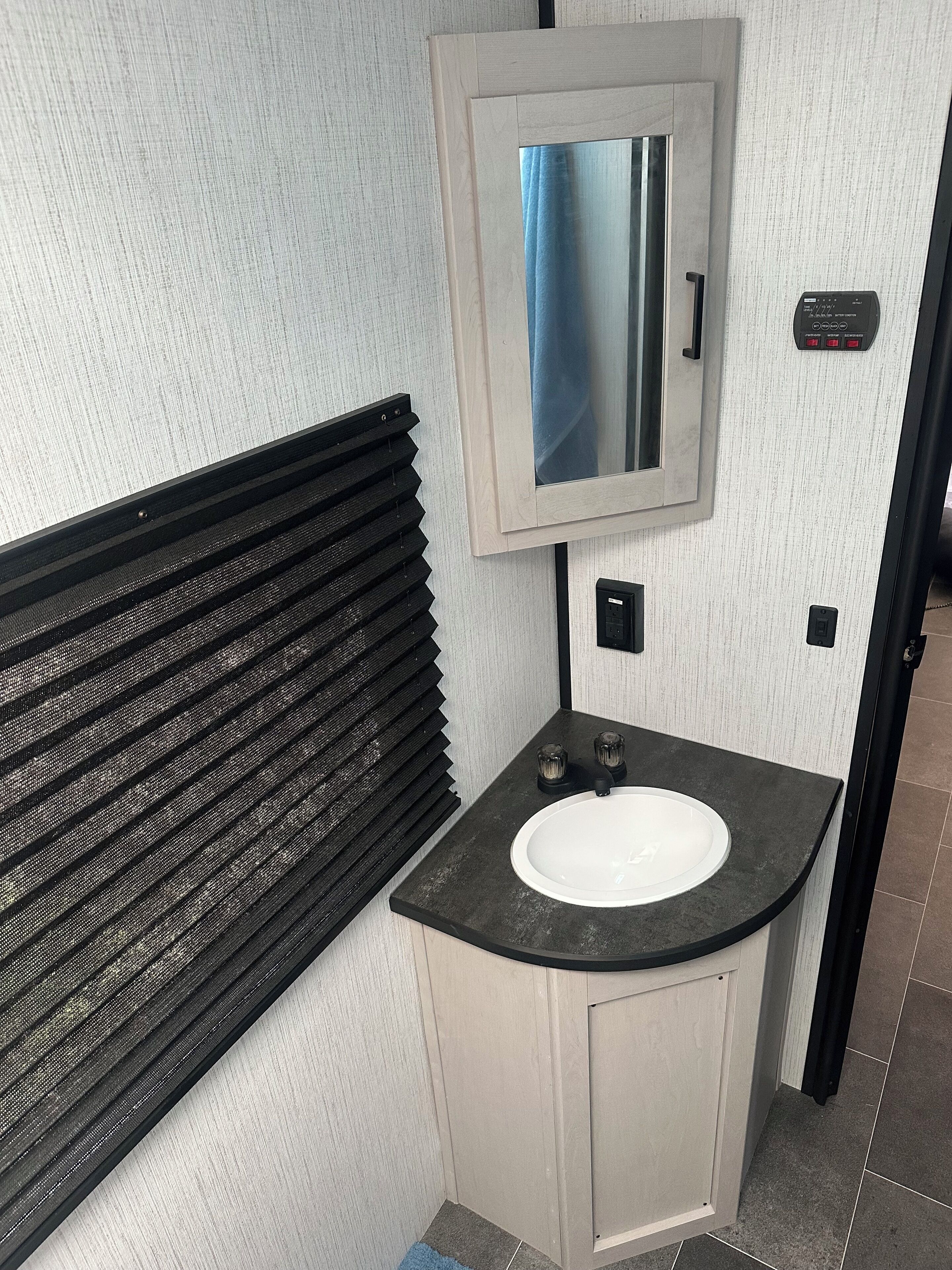 Ein Badezimmer mit einem weißen Waschbecken, einem Spiegel, einem schwarzen Lüftungsgitter und einem Lichtschalter.