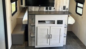 A compact RV interior with a flat-screen TV mounted above a kitchenette area, a small seating area, and a window for natural light.