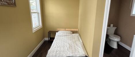 A small, narrow bedroom with a single bed, a window, and a door leading to a bathroom.