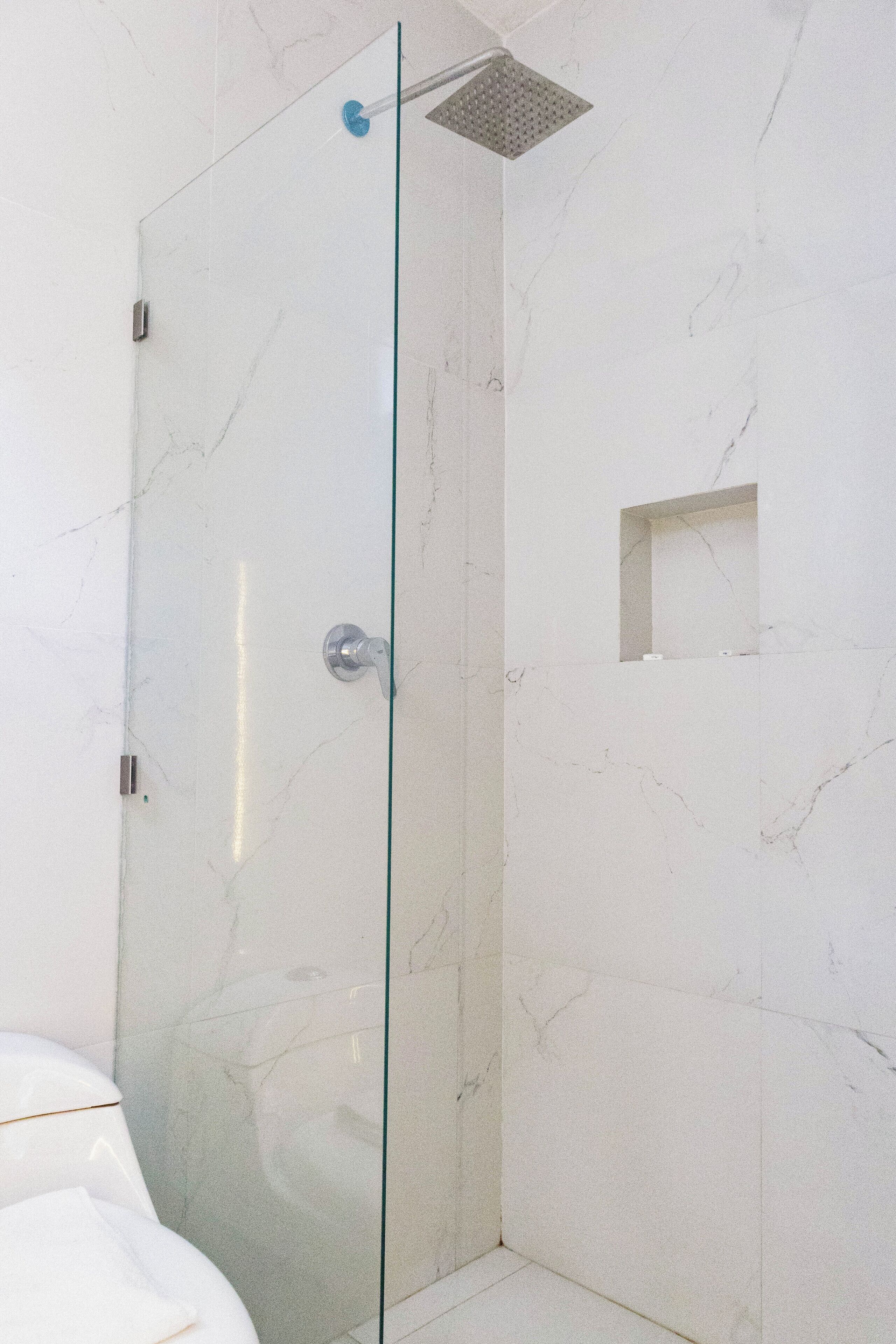 Une douche fermée par des parois de verre, dotée d’un pommeau de douche, d’une petite fenêtre et d’un mur recouvert de carreaux de marbre.