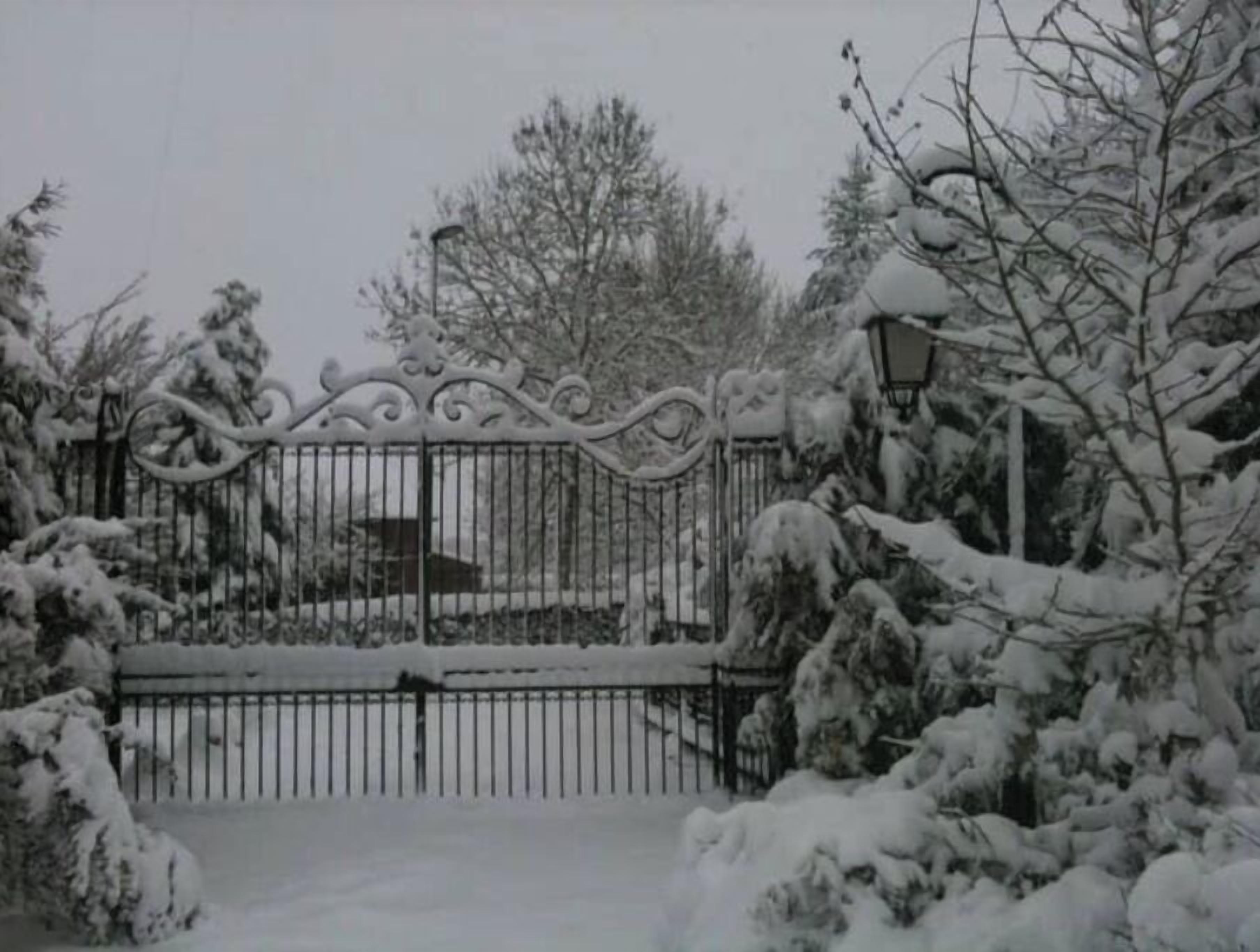 A snow-covered gate with intricate metalwork, leading to a building with a visible roof.