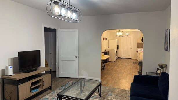 A living room with a TV, entertainment unit, glass coffee table, and a blue sofa. A kitchen is visible in the background with a dining area. - Charming 2-bdrm home in Little Rock with WiFi and AC (Little Rock)