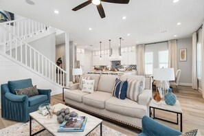 A modern living room with a sofa, coffee table, and blue armchairs. A staircase leads to an upper level. - Serenity Cove Beachside Village Galveston - Lux 4bd Sleeps 12 w/ Beach Access (Galveston)