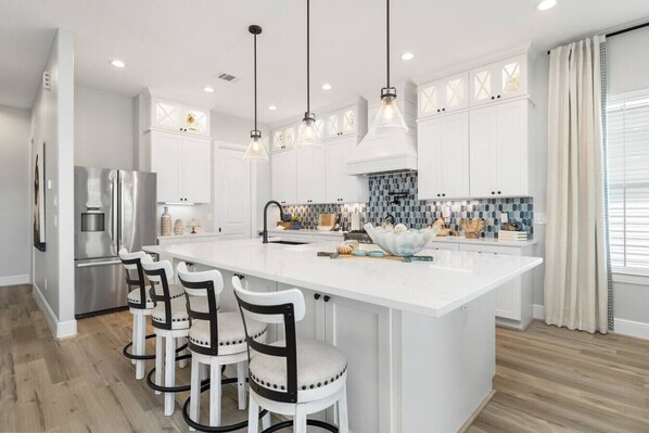 A modern kitchen with a large island, white cabinetry, stainless steel appliances, and pendant lighting. - Serenity Cove Beachside Village Galveston - Lux 4bd Sleeps 12 w/ Beach Access (Galveston)