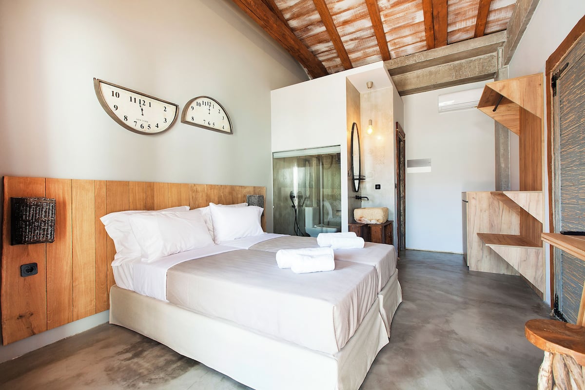 A bedroom with a wooden headboard, a large bed with white linens, two wall clocks, and a wooden staircase.