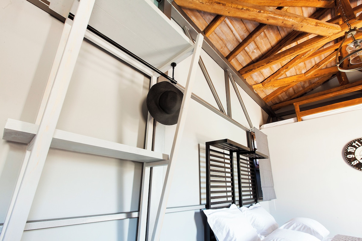 A room with a sloped ceiling, a bed with white bedding, a hat hanging on the wall, and a clock on the wall.