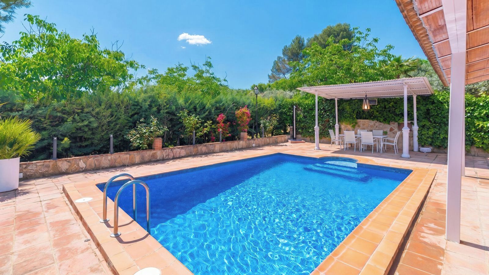 A rectangular swimming pool with a tiled edge, surrounded by a stone patio. There is a covered seating area with white chairs and a table.