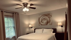 Ein Schlafzimmer mit einem Bett, zwei Nachttischlampen, einem Deckenventilator, einem Fenster mit Vorhängen und einem Wandbild einer Rose.