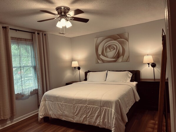 A bedroom with a bed, two bedside lamps, a ceiling fan, a window with curtains, and a wall-mounted rose painting. - The Cozy Gray House (Fort Mill)