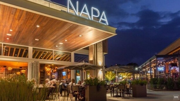 Ein Essbereich im Freien mit der Aufschrift 'NAPA', umgeben von Tischen und Stühlen, und ein überdachter Sitzbereich mit Sonnenschirmen.