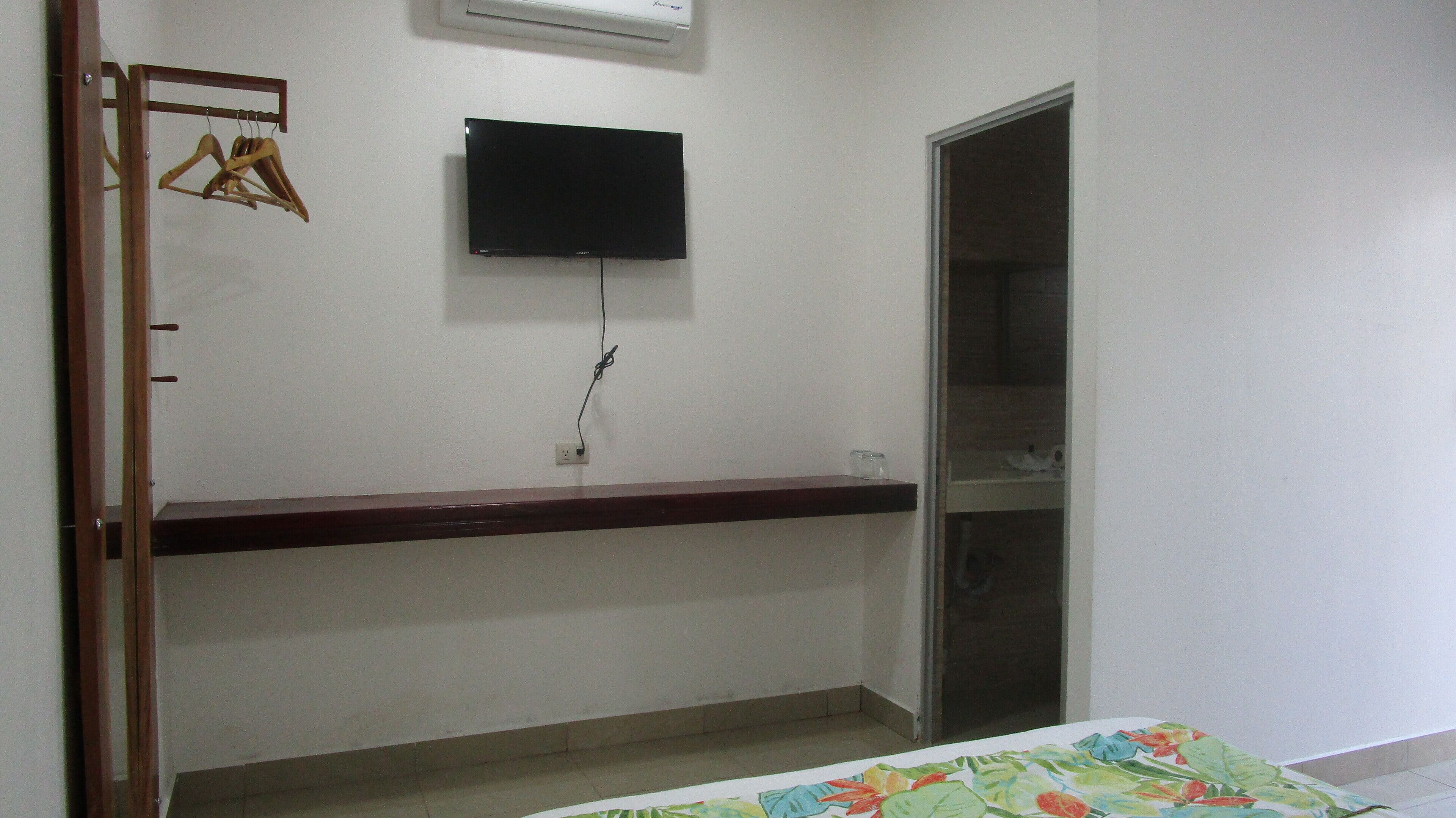 A bedroom with a bed, a TV mounted on the wall, a wooden hanger rack, and a door leading to a closet.