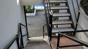 Una escalera con barandales de metal y un pasamanos, que conduce a un rellano con una pared blanca y vistas al exterior con vegetación.