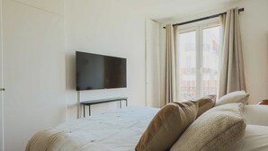 A bedroom with a bed, a TV mounted on the wall, a window with curtains, and a view of buildings outside.