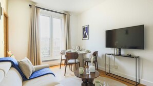 A modern living room with a sofa, a dining table set for four, a flat-screen TV on a stand, and a large window with curtains.