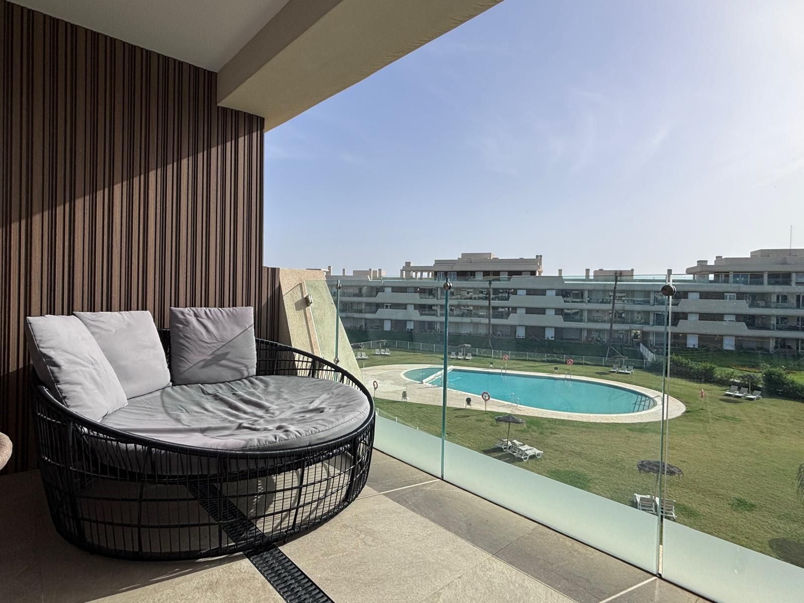 A balcony with a round black wicker chair and cushions, overlooking a swimming pool and a green lawn.