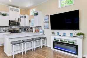 Une cuisine moderne avec des meubles blancs, un îlot central doté de tabourets, un four encastré et un grand téléviseur à écran plat.