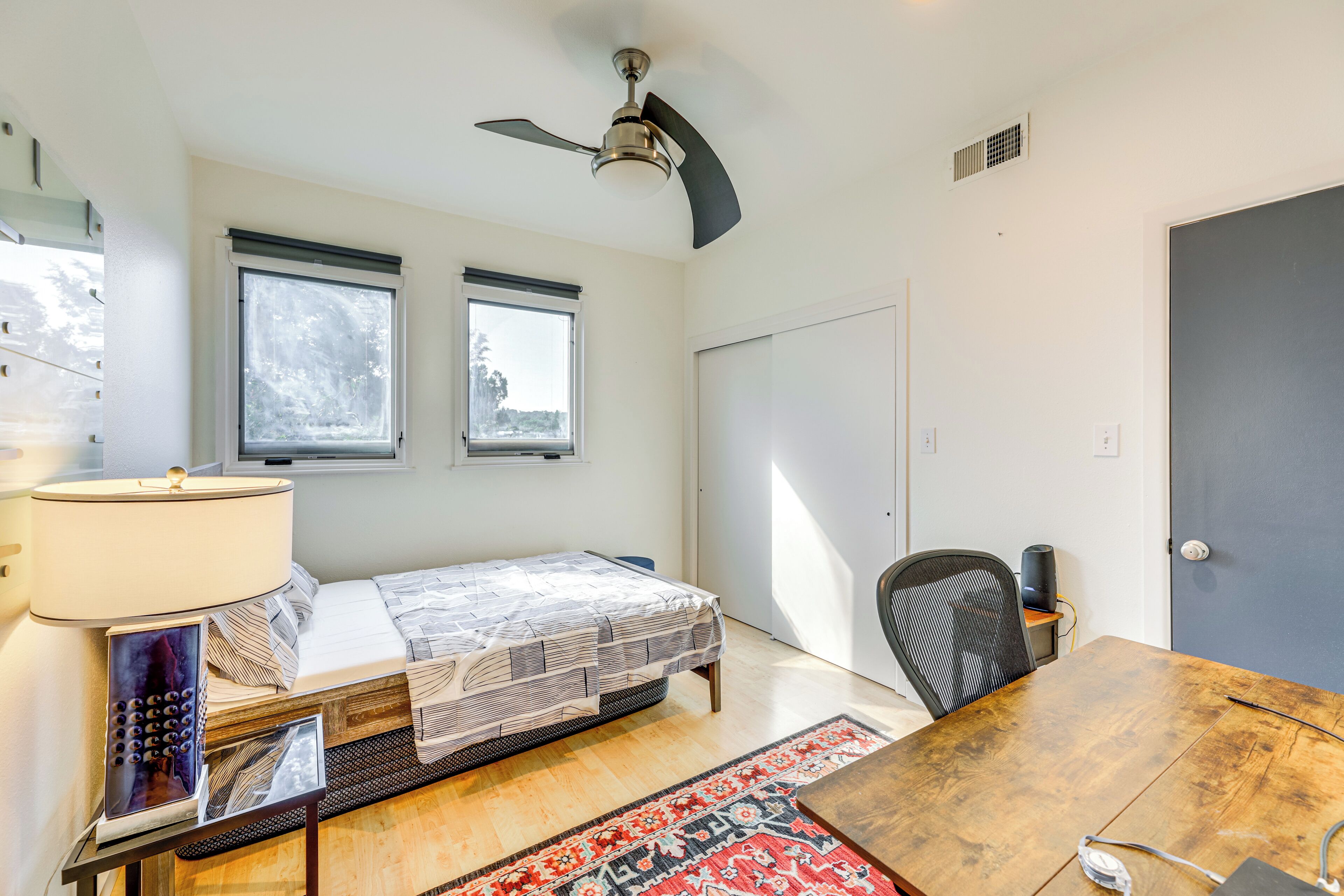 A bedroom with a bed, a desk, a chair, a ceiling fan, and two windows.