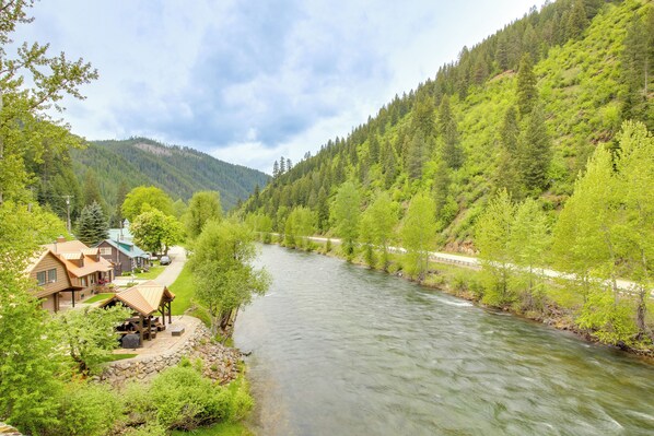 Una cabaña junto al río con terraza y asientos exteriores, rodeada de abundante vegetación y una ladera boscosa.
