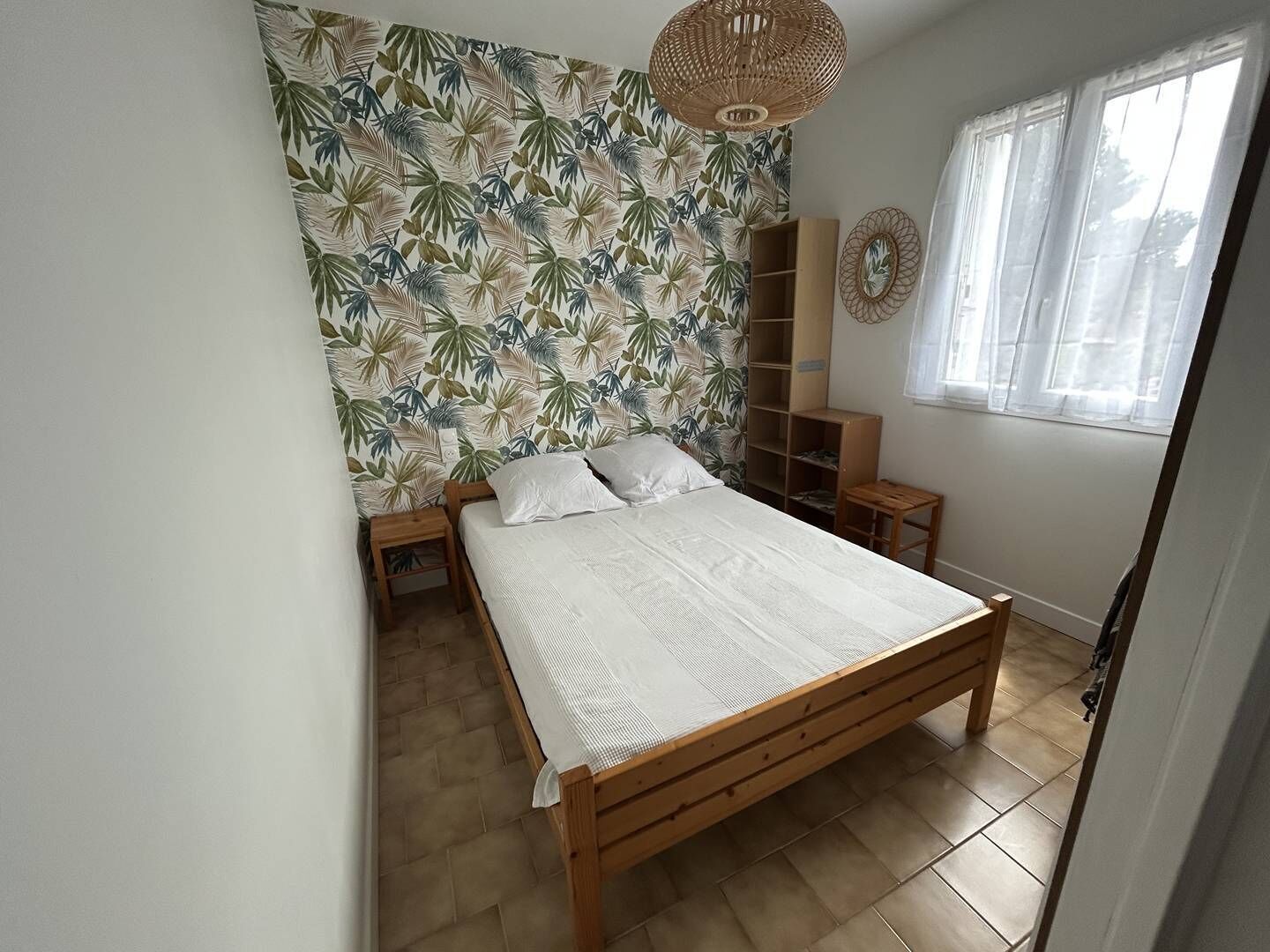 Quarto com cama de madeira, parede com padrão de folhagem tropical, luminária de palha no teto e janela com cortinas finas.