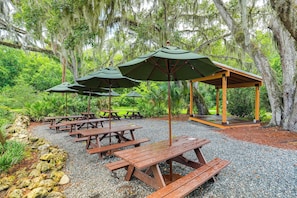 Un área de picnic cubierta con bancos de madera y sombrillas verdes, rodeada de abundante vegetación y árboles.
