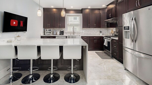 A modern kitchen with dark wood cabinets, stainless steel appliances, a central island with bar stools, and a flat-screen TV mounted on the wall. - New England Traditional x Modern, 5 Bedrooms, Office, 8ppl (Quincy)