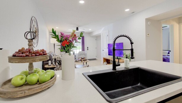 A modern kitchen with a white countertop, a black sink, a fruit display, and a vase with flowers.