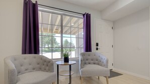 A room with two grey armchairs, a small round table with a potted plant, and a large window with purple curtains.