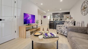 A modern living room with a flat-screen TV, a dining table with snacks, a sofa, and a kitchen area in the background.