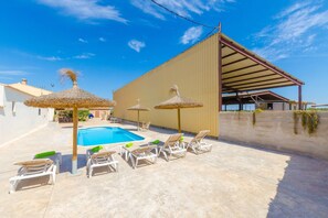 Uma área de piscina com espreguiçadeiras e guarda-sóis de palha, um edifício grande com telhado inclinado e um céu azul límpido.