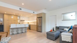 A modern kitchen with wooden cabinets, a stainless steel refrigerator, and a dining area with white chairs. - Modern Living Across from the Beach (Saanich North)