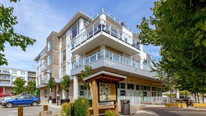 A modern multi-story building with a prominent address sign, outdoor seating, and a landscaped area with wooden posts and plants. - Modern Living Across from the Beach (Saanich North)