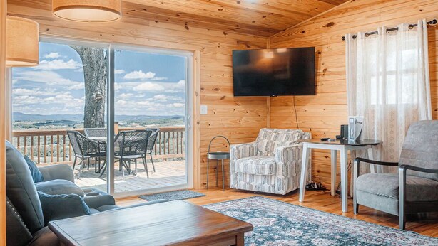 A cozy cabin interior with a wooden ceiling, a flat-screen TV, a sofa, a table, and a view through the glass door. - Pops Cozy Cabin w/ an incredible valley view. Gas firepit, full kitchen and more (Fancy Gap)