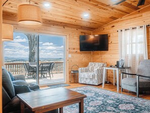 A cozy cabin interior with a wooden ceiling, a flat-screen TV, a sofa, a table, and a view through the glass door. - Pops Cozy Cabin w/ an incredible valley view. Gas firepit, full kitchen and more (Fancy Gap)